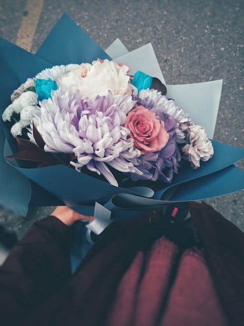A hand holding a bouquet of fresh flowers wrapped in dark blue paper, featuring a large light purple chrysanthemum at the center, complemented by a pale pink rose, white and purple carnations, and hin