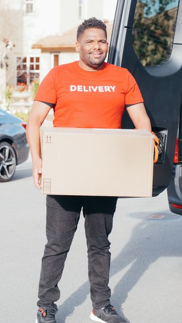 A smiling male delivery driver with short curly hair and a beard, wearing a bright orange T-shirt with 'DELIVERY' printed in white, stands outdoors near a black delivery van. He holds a large, plain c