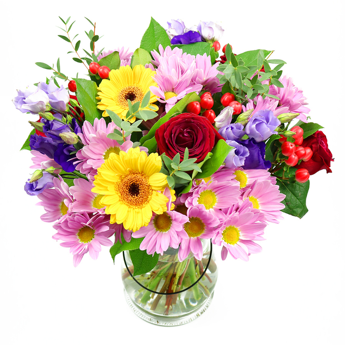 In this image, a lush, overflowing bouquet commands attention from the mouth of a clear glass vase, placed against a crisp white backdrop that makes every colour glow. At the centre, deep red roses open in generous spirals, their velvety petals hinting at whispered sentiments and long-held affection. Radiating out from them, bright yellow gerbera daisies with distinctive brown-orange centres create a ring of sunshine, lending the whole piece a mood of cheerful celebration. Embracing these focal blooms is a halo of pink daisies or chrysanthemums, their many slender petals and small yellow eyes forming soft, cushiony clusters that add both volume and tenderness. Slender stems of lisianthus in shades of purple and lavender weave through the design, their bell-shaped flowers introducing graceful curves and a cool, evening-sky tone that balances the warmth of the gerberas and roses. Scattered throughout are glossy red hypericum berries, perfectly round and jewel-like, offering a textural contrast that feels modern and slightly playful. An abundance of greenery frames the flowers: broad, dark leaves anchor the design, while lighter green sprigs and shoots add lift and a just-picked freshness, reminiscent of a stroll through a hidden garden off the streets of Canning Town or near the Royal Victoria Dock. The glass vase reveals the neatly cut stems resting in crystal-clear water, evidence of the care taken in conditioning each flower. Overall, the bouquet feels like a vibrant, uplifting gift-ideal as a dining table centrepiece in an E16 apartment, a thank-you after a gathering at a local café, or a gesture of congratulations that brings a burst of colour and life indoors.